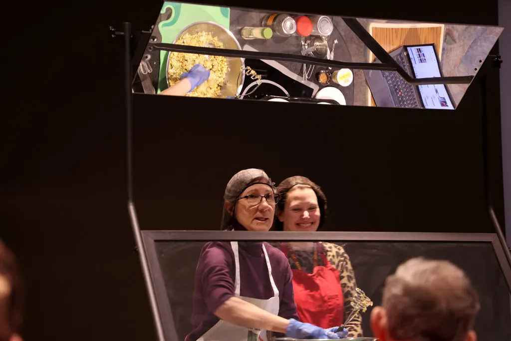Two women in aprons and hairnets smiling together at a cooking demonstration, with an overhead mirror showing their hands mixing food in a bowl