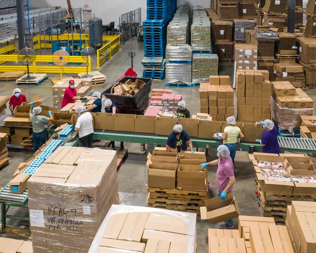 Overhead view of volunteers in hairnets and gloves packing food into cardboard boxes along a warehouse assembly line